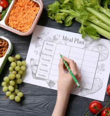 Photograph of a hand and pencil completing a meal planner with fruit and containers of food around edges of image