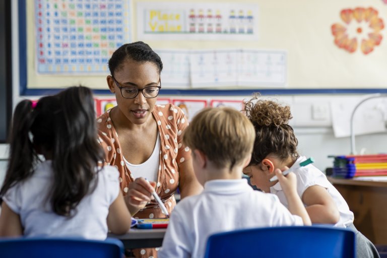 A teacher works with a small group of primary school children gathered around a table, helping them with a classroom learning activity.
