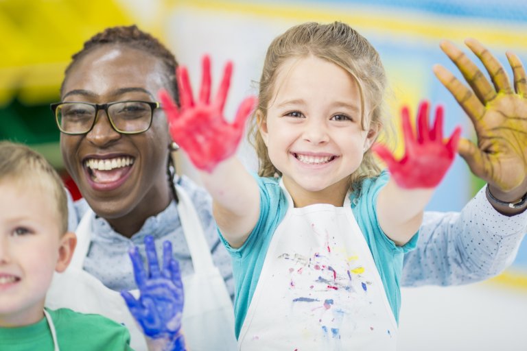 Smiling children and an adult take part in a painting activity, holding up colourful paint-covered hands.
