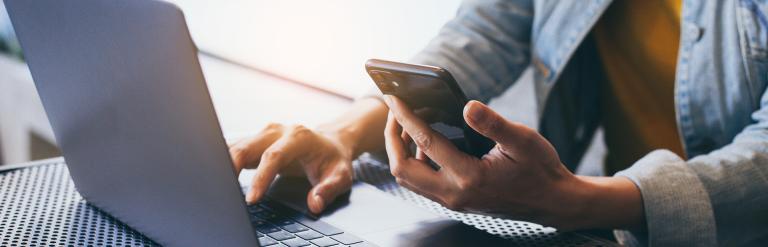 A person looking at a laptop and holding a phone.