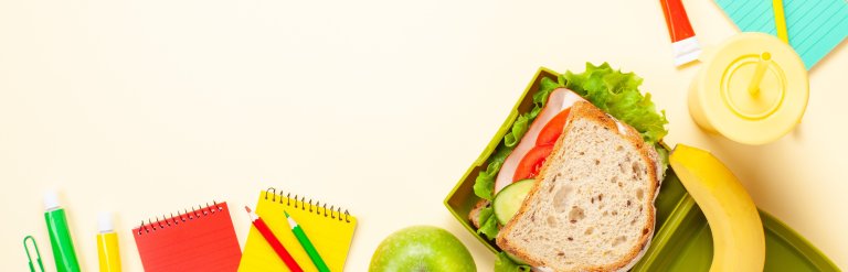 Photograph of an open lunchbox surrounded by healthy food items and colourful stationary against a cream background