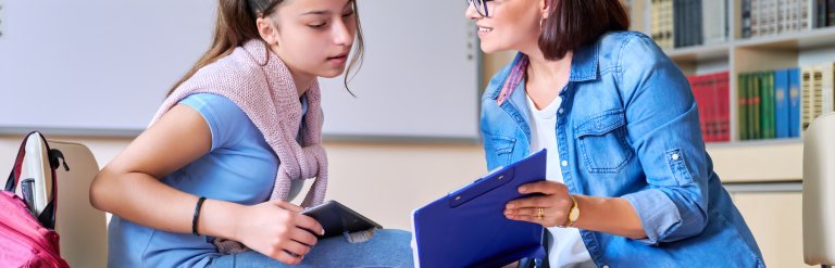 A teacher or support worker talks with a teenage student in a school library or classroom, offering one-to-one guidance while reviewing notes on a clipboard.
