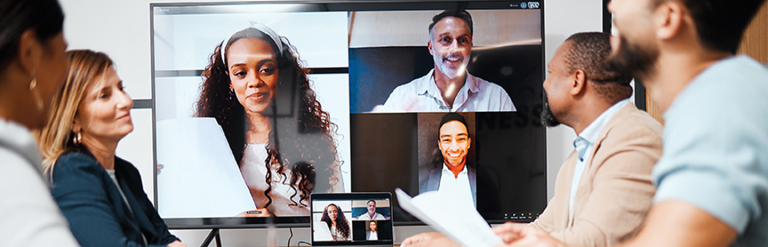 A photograph of a meeting with four individuals sat at a table and three more connected via a video conference call
